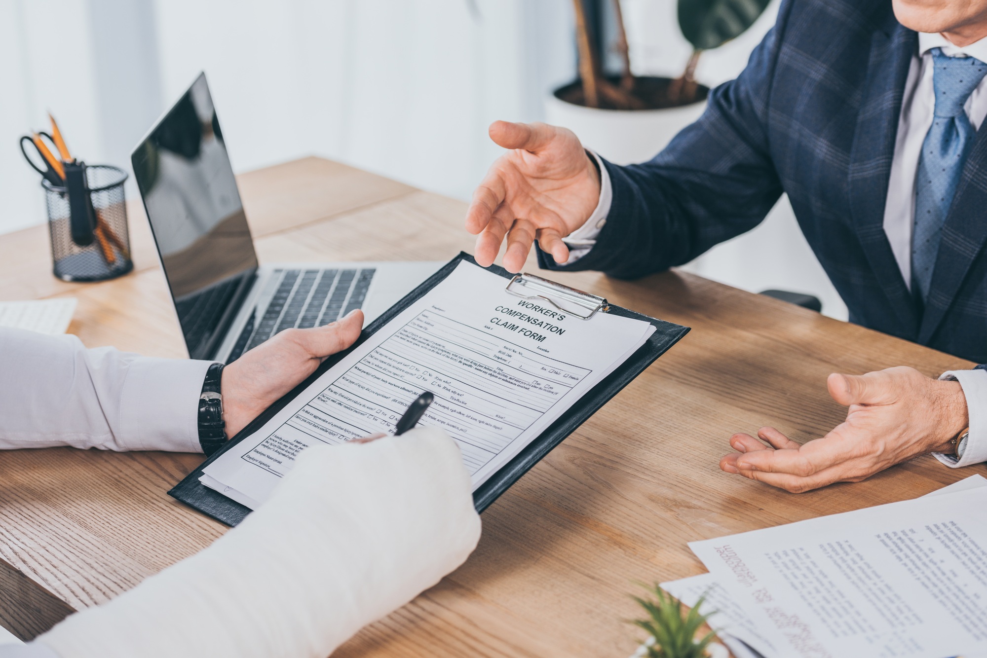 cropped view of worker with broken arm signing form for compensation claim opposite to businessman