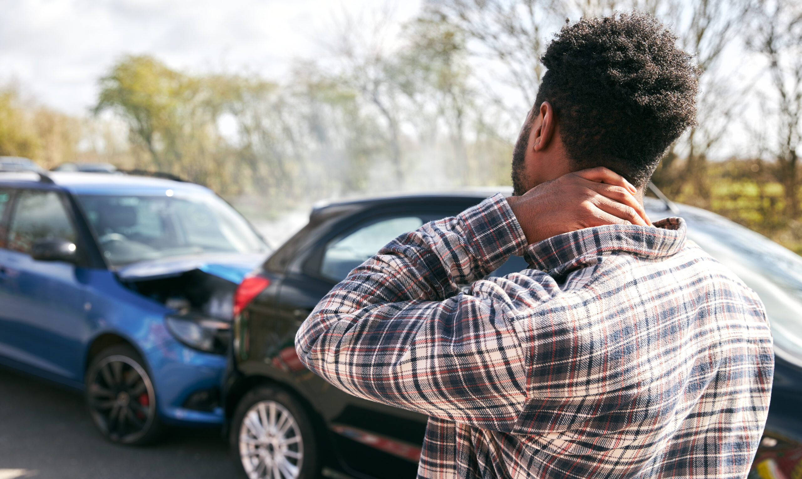 Young Male Driver With Whiplash Injury Standing By Damaged Car After Road Traffic Accident